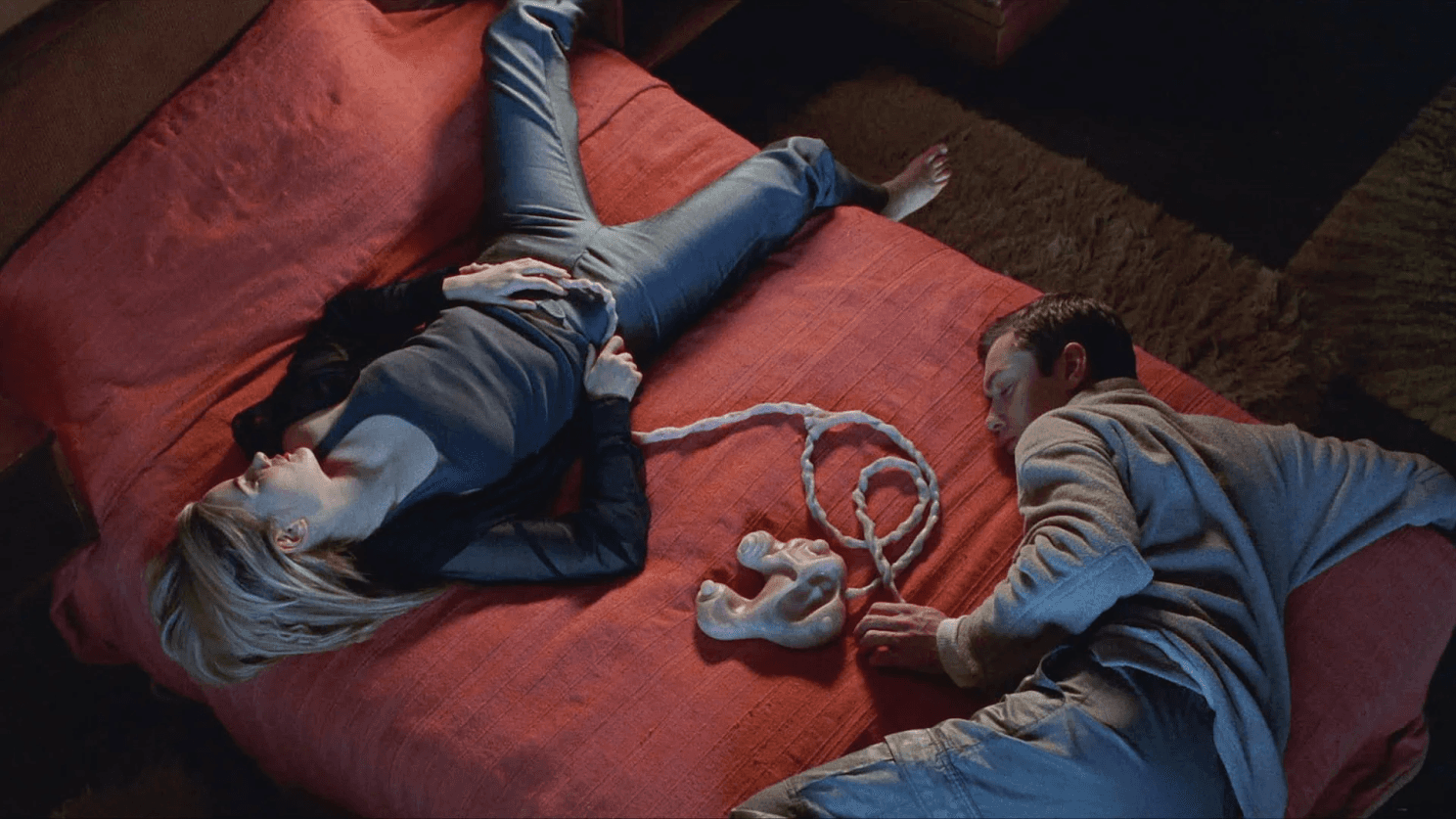 Plan aérien de deux personnages allongés sur un lit rouge, reliés par un cordon organique à une manette de jeu bio-mécanique, scène du film eXistenZ
