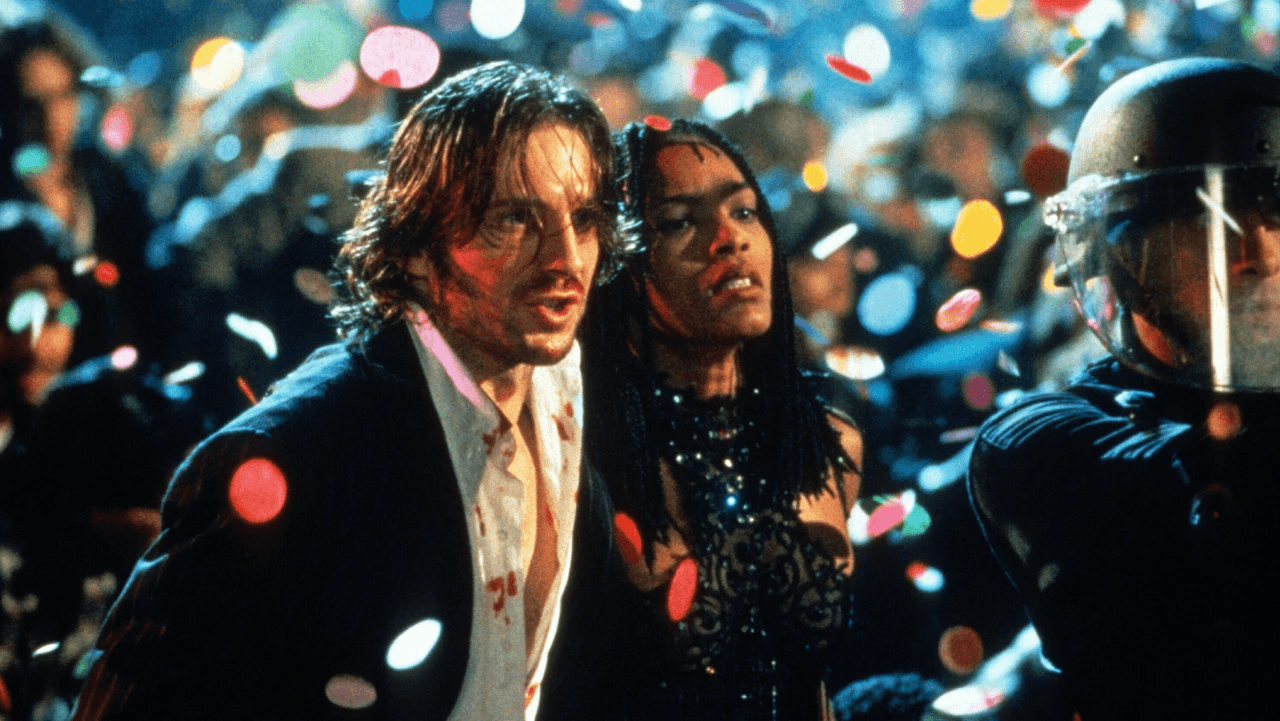 Deux personnages au premier plan dans une foule en liesse sous des confettis, un policier casqué à leurs côtés, scène du film Strange Days